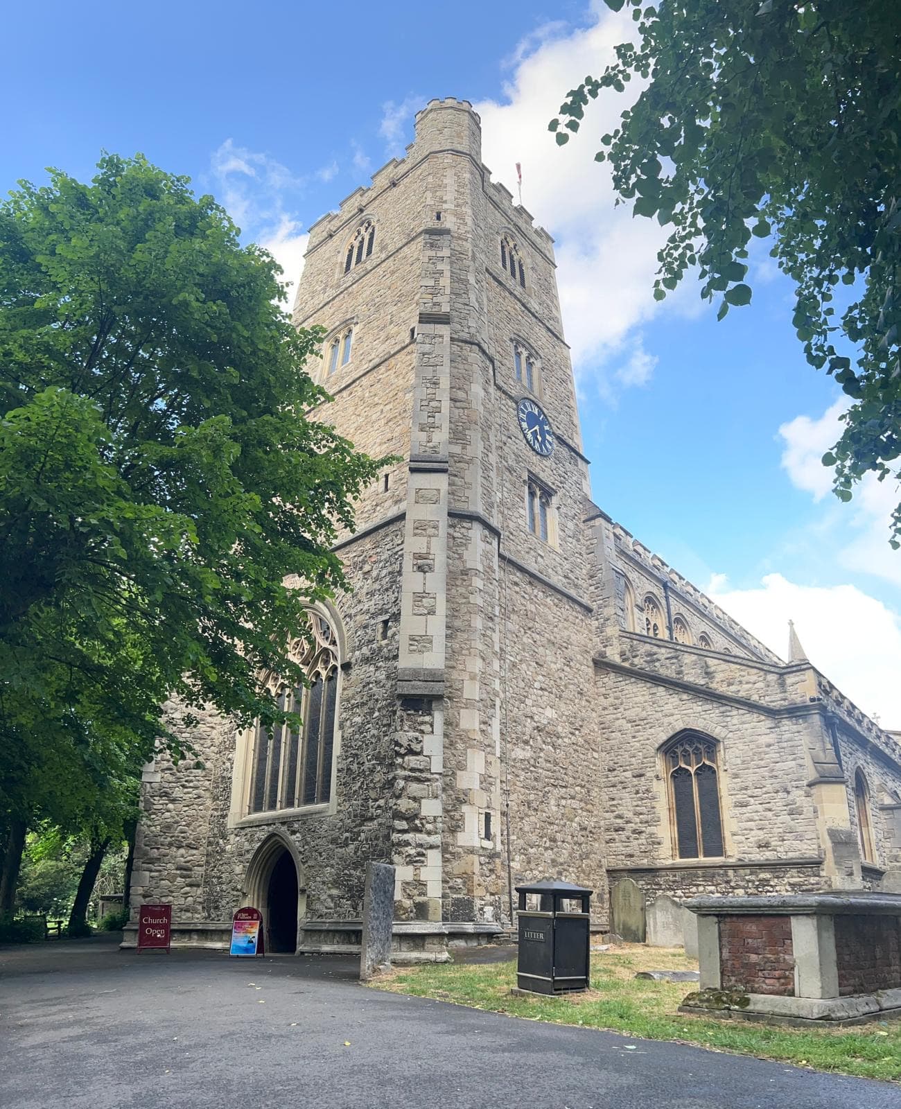 Stone tower of All Saints Church, Fulham, with a clock face, framed by green trees