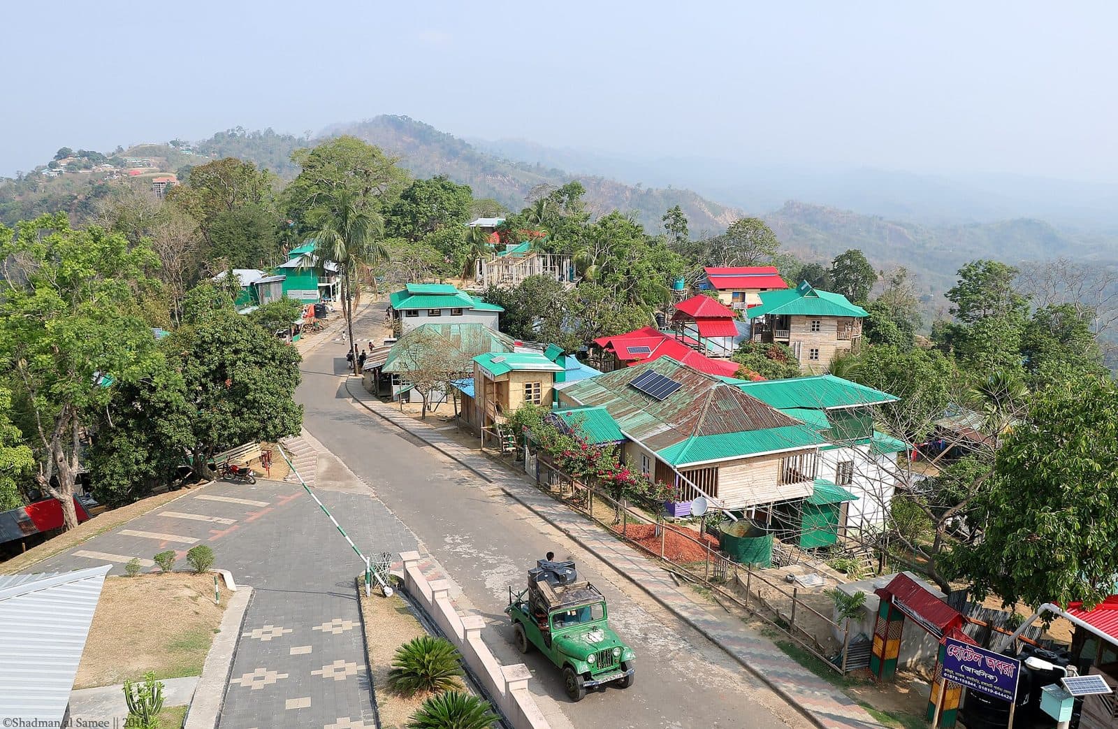 Colourful cottages and a jeep on the main road through Ruilui Para in Sajek Valley, with green hills behind