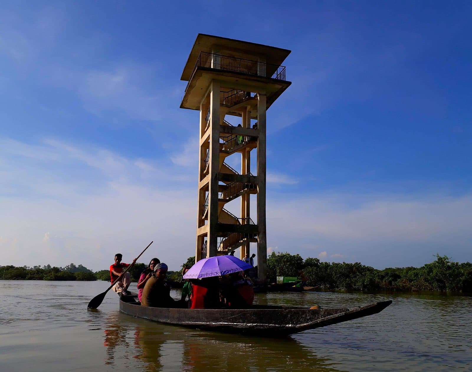 Wooden boat with visitors passing a tall watchtower on the water at Ratargul Swamp Forest